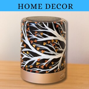 May include: A black and white speaker with a gold base. The speaker is decorated with a white tree branch design and orange and gold leaves. The speaker is a home decor item.