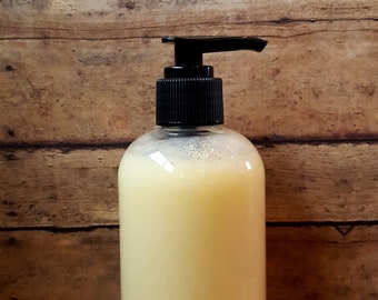 May include: A clear pump bottle with a black pump top filled with a light yellow liquid. The bottle is sitting on a wooden surface.