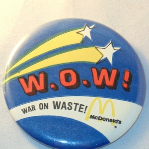 Puede incluir: Un botón azul y amarillo con el texto "W.O.W! War on Waste!" y el logotipo de McDonald's.