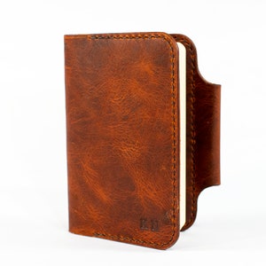 May include: A brown leather notebook cover with a stitched edge and a pen loop. The cover has a distressed look and the initials "K.M." are embossed on the front.