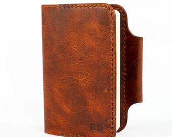 May include: A brown leather notebook cover with a stitched edge and a pen loop. The cover has a distressed look and the initials "K.M." are embossed on the front.