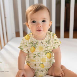 May include: A baby wearing a yellow and white bodysuit with a lemon and bee print. The bodysuit has a gingham pattern and features watercolor illustrations of lemons, leaves, and bees.