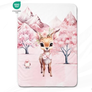 Peut inclure: Une illustration aquarelle d'un faon avec un nœud rose et une couronne de fleurs se tenant dans un paysage enneigé rose et blanc avec deux cerisiers en fleurs roses. Le texte "Oeko-Tex Standard 100" est dans le coin supérieur gauche de l'image.