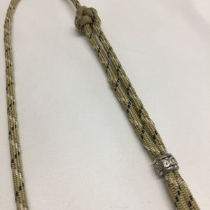 May include: A tan and black paracord lanyard with a silver bead. The bead has an inscription on it.