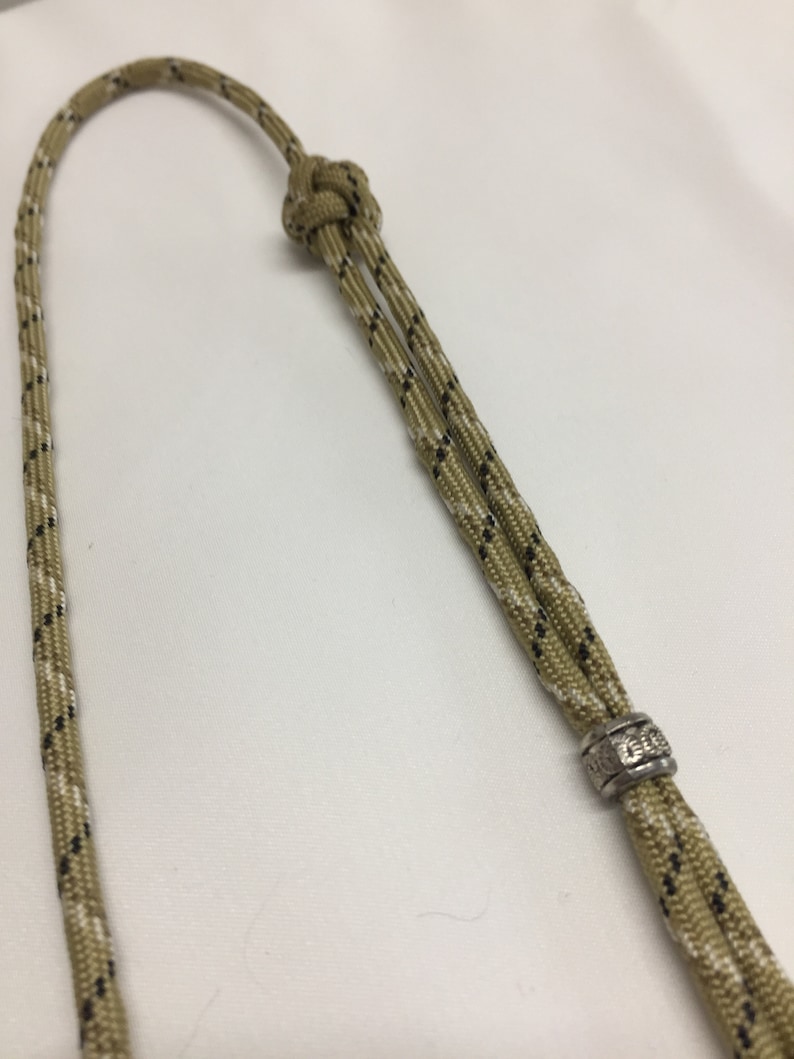 May include: A tan and black paracord lanyard with a silver bead. The bead has an inscription on it.