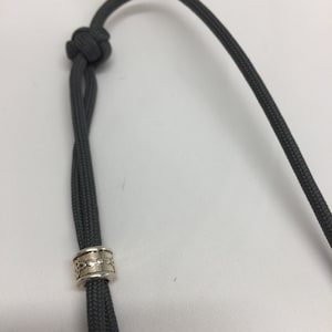 May include: A gray paracord lanyard with a silver bead. The bead has a decorative pattern.