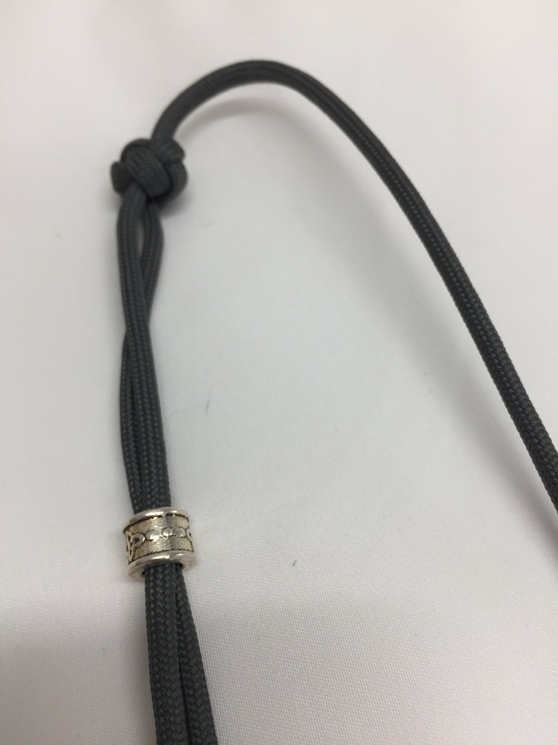 May include: A gray paracord lanyard with a silver bead. The bead has a decorative pattern.