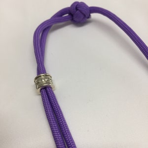May include: A purple paracord lanyard with a silver bead. The lanyard is tied with a knot at the top.