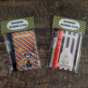 May include: Two Halloween notebook and pen sets in clear plastic packaging. The left notebook features orange and black diagonal stripes with a black cat graphic. The right notebook has black and white vertical stripes with a ghost graphic.