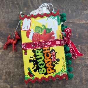 May include: A Sour Patch Kids candy box decorated for the holidays with a red ribbon printed with the text NO PEEKING, green pom-pom trim, red rickrack trim, a red glittery reindeer ornament, and a gift tag with the text To: From:.