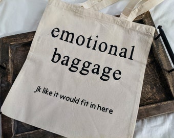 May include: A white canvas tote bag with black text that reads "emotional baggage jk like it would fit in here". The bag is sitting on a wooden tray.
