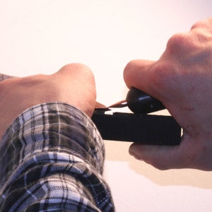 May include: A person's hand using a black and silver tool to cut a piece of black foam.