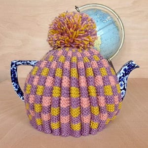 May include: A purple and yellow knitted teapot cosy with a fluffy pom-pom on top. The cosy is covering a blue and white teapot.