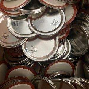 May include: A pile of silver metal jar lids with red rubber seals. The lids are labeled "Ball Sure Tight".