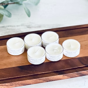 Könnte beinhalten: Sechs kleine, weiße Teelichter mit dem Text "London James Candle Co." auf einem Holzschneidebrett. Die Kerzen sind mit verschiedenen Düften parfümiert, darunter Pfefferminze, Eukalyptus und Grapefruit. Das Schneidebrett ist aus hellem und dunklem Holz.