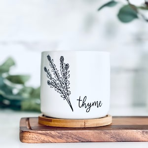 Pode incluir: Vaso de cerâmica branco com um desenho de linha preta de um ramo de tomilho e a palavra "tomilho" escrita em caligrafia preta. O vaso está sobre uma superfície de madeira.