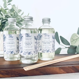 Op de afbeelding: Drie doorzichtige glazen flessen met zilveren deksels en etiketten. De etiketten zijn wit met blauwe bloemmotieven en tekst. De flessen zijn gevuld met een heldere vloeistof en zijn gelabeld met "Library", "French Linen" en "Sea Salt & Orchid". De flessen staan op een houten oppervlak met houten stokjes.