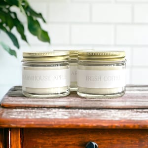 Puede incluir: Tres tarros de velas de vidrio con tapas doradas y etiquetas blancas. Las etiquetas dicen "Farmhouse Apple", "Fresh Coffee" y "Vanilla Bean".