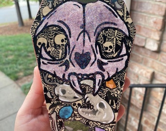 May include: A coffin-shaped wooden box decorated with a glittery cat skull, bones, and other spooky imagery. The skull has a heart shape in the center and a witch's hat on top.