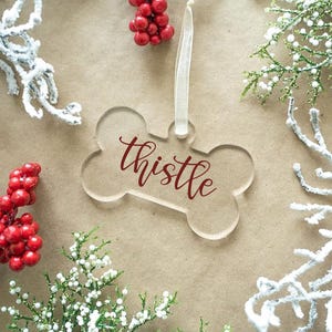May include: A clear acrylic bone-shaped ornament with the word "thistle" in burgundy script. The ornament hangs from a white ribbon. The background features red berries and white frosted branches, suggesting a holiday theme.
