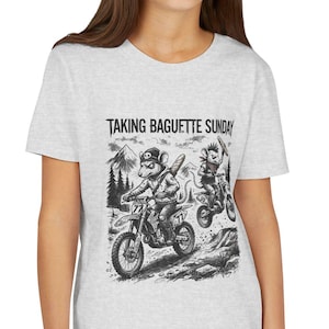May include: A light gray t-shirt featuring a black and white graphic of cartoon characters riding motorcycles. The text "TAKING BAGUETTE SUNDAY" is printed above the graphic. The shirt is being worn by a person.