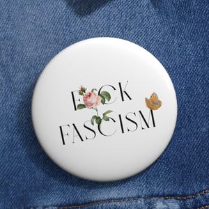 Könnte beinhalten: Weißer Knopf mit dem Text "F*CK FASCISM" in schwarzen Buchstaben. Auf dem Knopf sind eine rosa Rose und ein Schmetterling abgebildet.