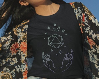 May include: A woman wearing a black t-shirt with a white outline of a 20-sided die, a crescent moon, and stars. The die is surrounded by two hands. She is wearing a floral kimono and denim shorts.