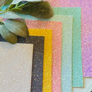 May include: A set of eight glitter cardstock sheets in various colors, including white, black, gold, pink, light blue, and purple. The sheets are arranged in a fan shape, with the white sheet at the bottom and the purple sheet at the top.