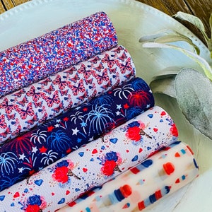 May include: A collection of rolled faux leather sheets with patriotic designs. Patterns include red, white, and blue glitter, butterflies, fireworks, ice cream cones, and scattered stars and hearts. The sheets are arranged on a white, oval-shaped tray.