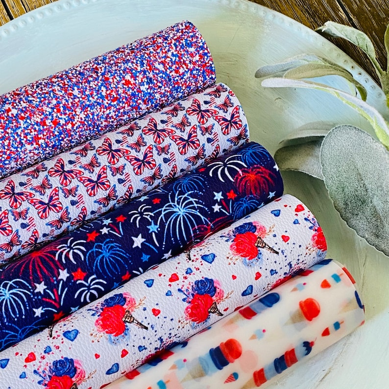 May include: A collection of rolled faux leather sheets with patriotic designs. Patterns include red, white, and blue glitter, butterflies, fireworks, ice cream cones, and scattered stars and hearts. The sheets are arranged on a white, oval-shaped tray.