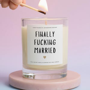 May include: A lit candle in a clear glass jar with the text "FINALLY FUCKING MARRIED" on a white label. The candle sits on a pink circular stand. The candle is being lit by a match.