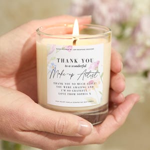 May include: A burning vanilla-scented soy wax candle in a clear glass. The candle features a label with the text "THANK YOU to a wonderful Make-up Artist" and a floral design. The candle is held in two hands.