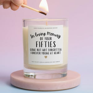 May include: A clear glass candle with a white label featuring black text that reads In loving memory of your fifties, gone but not forgotten, forever young at heart, with a small gold heart icon and rich velvet vanilla scented soy wax.