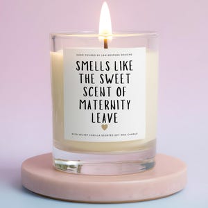 May include: A lit soy wax candle in a clear glass jar with a white label that reads: HAND POURED BY L&M BESPOKE DESIGNS, SMELLS LIKE THE SWEET SCENT OF MATERNITY LEAVE, with a small heart icon and RICH VELVET VANILLA SCENTED SOY WAX CANDLE.