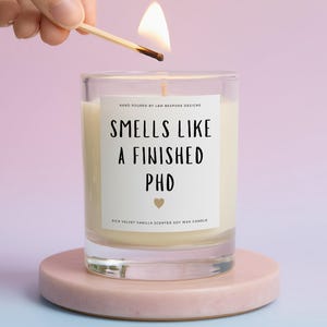 May include: A clear glass candle with a white label that reads "SMELLS LIKE A FINISHED PHD" in black text, featuring a small gold heart icon. The candle contains off-white wax and is being lit by a wooden matchstick, emitting a bright flame.