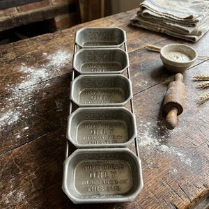 May include: A metal bread tin rack with five individual loaf pans. Each pan is inscribed with "HOVIS BREAD ONLY TO BE BAKED IN THIS TIN". The rack sits on a rustic wooden surface, with baking supplies in the background.
