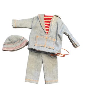 May include: A doll-sized outfit consisting of a light blue long-sleeved hooded jacket with a red and white striped lining, matching long pants with orange stitching, and a small matching hat with red circular stitching details on the brim.