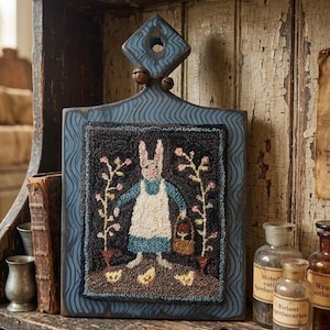 May include: A blue wooden decorative cutting board with a rug-hooked design. The rug features a bunny wearing a blue dress and white apron, holding a basket. The board has a diamond-shaped handle with two small bells.