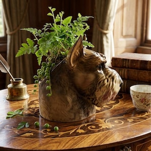 May include: A brown ceramic dog head planter filled with green plants and trailing vines. The planter sits on a wooden table with inlaid floral designs, next to a brass inkwell and a teacup.