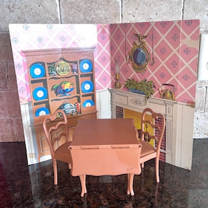 May include: A vintage Barbie doll dining set featuring a table, two chairs, and a backdrop of a pink wallpapered room with a cabinet and fireplace. The table and chairs are a light brown color.