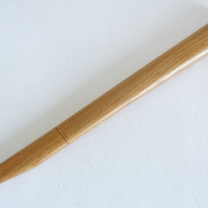 Op de afbeelding: Een lange, dunne houten pen met een licht afgerond uiteinde. De pen is lichtbruin van kleur en heeft een gladde, gepolijste afwerking.