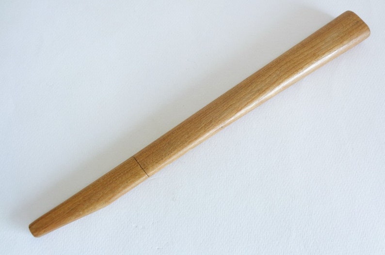 Op de afbeelding: Een lange, dunne houten pen met een licht afgerond uiteinde. De pen is lichtbruin van kleur en heeft een gladde, gepolijste afwerking.
