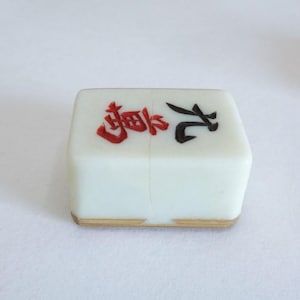 May include: A white Mahjong tile with red and black Chinese characters. The tile has a light brown edge and is set against a white background. This tile is a component of the classic game of Mahjong.