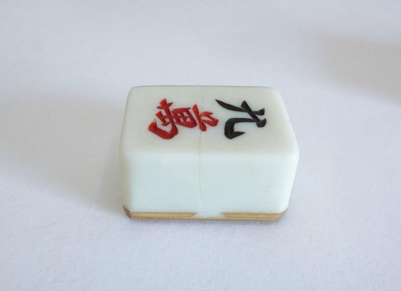 May include: A white Mahjong tile with red and black Chinese characters. The tile has a light brown edge and is set against a white background. This tile is a component of the classic game of Mahjong.