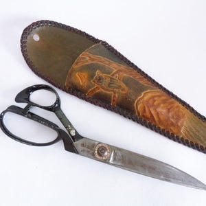 May include: A pair of vintage scissors with a black handle and silver blades, next to a leather sheath. The sheath is olive green with a brown, embossed design of a bird and tree. The sheath is approximately 10 inches long.