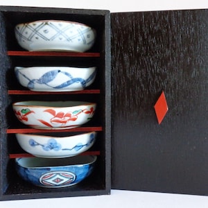Puede incluir: Un juego de cinco cuencos pequeños, redondos, de porcelana con varios diseños pintados en azul y rojo, anidados dentro de una caja de madera negra con una incrustación de diamante rojo.
