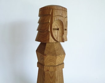 May include: A wooden sculpture of a stylized human figure with a geometric head and body. The sculpture is made of light brown wood and has a smooth, polished finish.