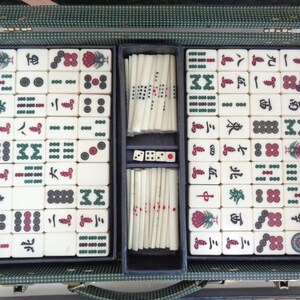 May include: Open vintage Mahjong set in a green case. The set includes ivory-colored tiles with red, green, and black symbols and numbers. The case is divided into compartments for tiles, dice, and sticks.