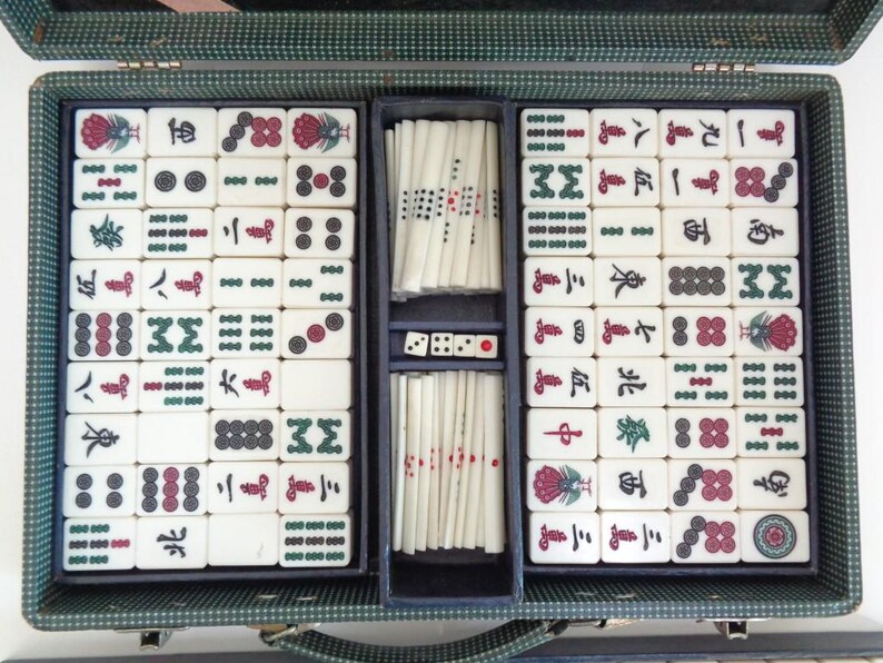 May include: Open vintage Mahjong set in a green case. The set includes ivory-colored tiles with red, green, and black symbols and numbers. The case is divided into compartments for tiles, dice, and sticks.