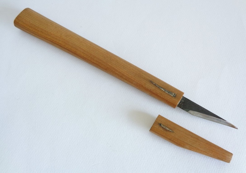Op de afbeelding: Een houten meshendel met een metalen blad. De hendel is gemaakt van lichtbruin hout en heeft een metalen inleg. Het blad is dun en scherp. Het mes is gescheiden van de hendel, waardoor de twee delen zichtbaar zijn.